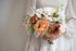 A close-up of a person in a white dress holding a rustic-style floral bouquet with peach, white, and brown flowers and green foliage.