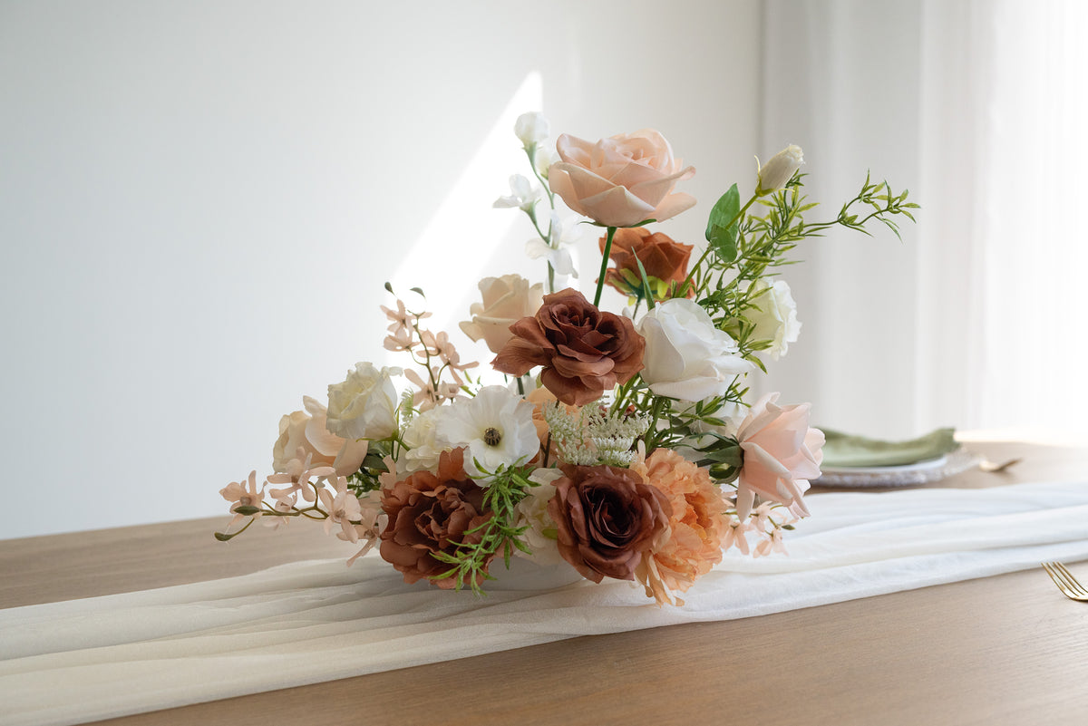A rustic floral centerpiece with peach, brown, and white flowers arranged on a white table runner over a light wood table.