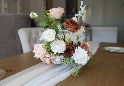 A floral centerpiece arrangement with white, blush pink, and brown roses, green foliage, and a sheer white runner on a wooden table in a softly blurred indoor setting.
