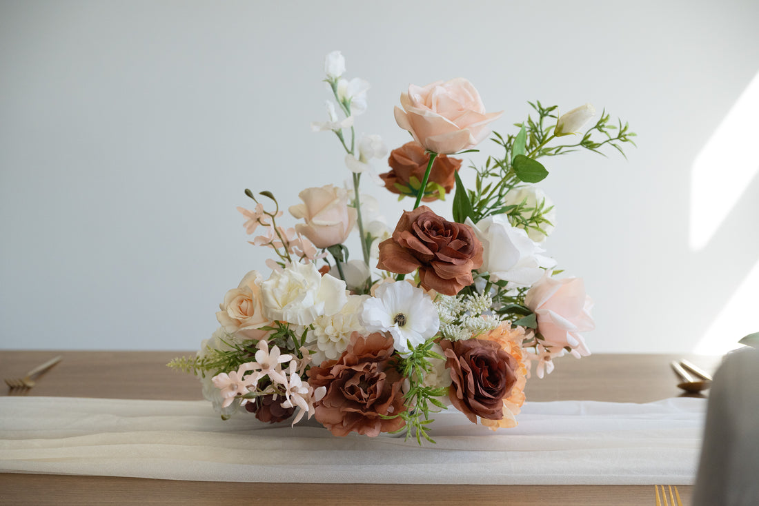 A floral centerpiece arrangement featuring blush pink, white, and warm brown faux roses with green foliage, displayed on a table runner against a light neutral background.