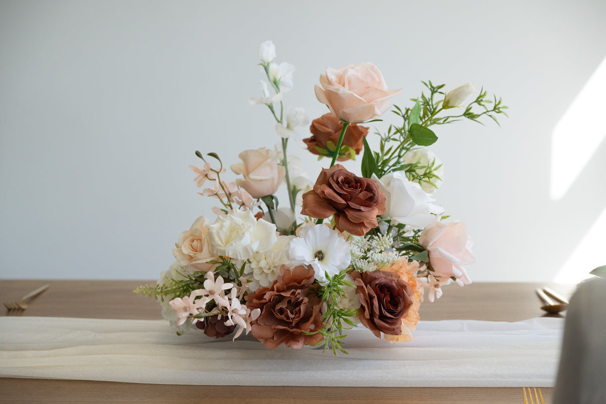 A floral centerpiece arrangement featuring blush pink, white, and warm brown faux roses with green foliage, displayed on a table runner against a light neutral background.