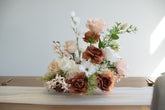 A floral centerpiece arrangement featuring blush pink, white, and warm brown faux roses with green foliage, displayed on a table runner against a light neutral background.