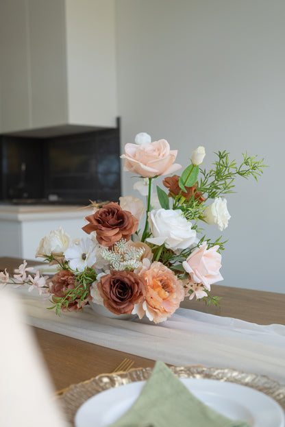 A rustic floral centerpiece with blush pink, terracotta, and white flowers arranged on a white tulle runner over a wooden table, with a blurred dining setting in the background.