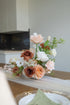 A rustic floral centerpiece with blush pink, terracotta, and white flowers arranged on a white tulle runner over a wooden table, with a blurred dining setting in the background.