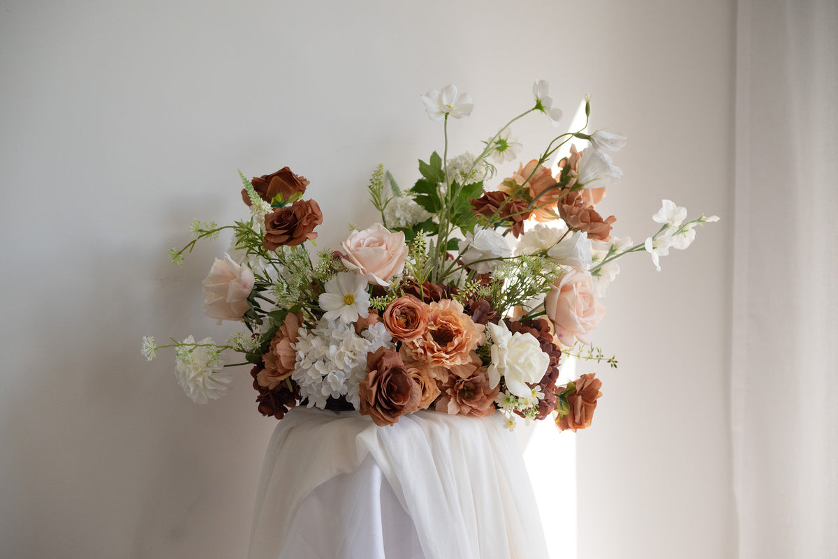 A large artificial floral arrangement featuring blush pink, terracotta, and white flowers with green foliage, displayed on a draped white table against a neutral off-white wall.