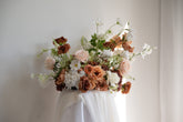 A large artificial floral arrangement featuring blush pink, terracotta, and white flowers with green foliage, displayed on a draped white table against a neutral off-white wall.