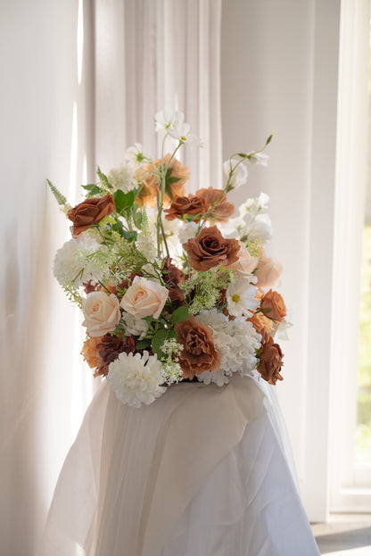 A large, lush floral arrangement featuring white roses, peach roses, and white daisies with green foliage, draped over a white fabric-covered stand in front of a softly lit window with sheer curtains.