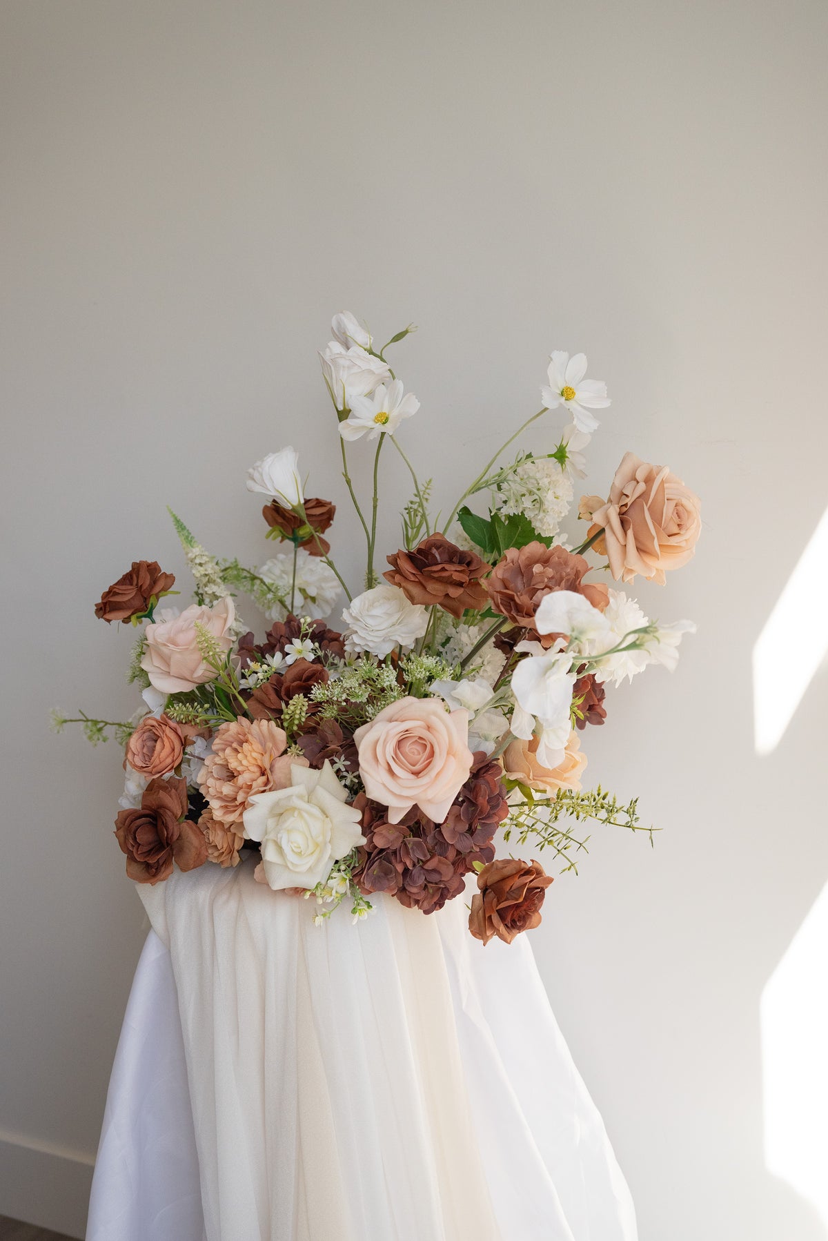 A rustic floral arrangement featuring blush pink, terracotta, and white flowers with green foliage, draped over a white fabric-covered stand against a neutral light gray wall.