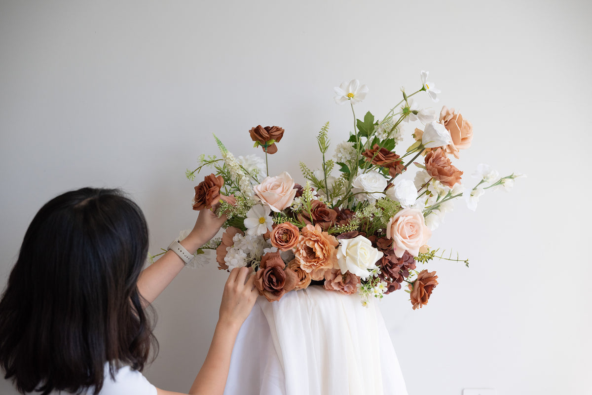 A woman with dark hair, seen from behind, adjusting a large artificial floral arrangement. The bouquet features a mix of dusty pink, terracotta, white, and green foliage, displayed against a plain white wall.