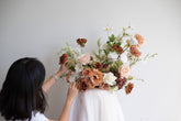A woman with dark hair, seen from behind, adjusting a large artificial floral arrangement. The bouquet features a mix of dusty pink, terracotta, white, and green foliage, displayed against a plain white wall.