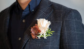 A close-up of a person wearing a dark gray plaid blazer over a navy button-down shirt, featuring a soft peach rose and brown flowers in a boutonniere on the chest pocket.
