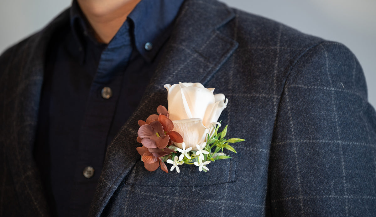 A close-up of a person wearing a dark gray plaid blazer over a navy button-down shirt, featuring a soft peach rose and brown flowers in a boutonniere on the chest pocket.