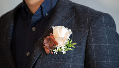 A close-up of a person wearing a dark gray plaid blazer over a navy button-down shirt, featuring a soft peach rose and brown flowers in a boutonniere on the chest pocket.