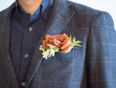 A close-up of a man wearing a dark gray plaid suit jacket over a navy blue button-down shirt, featuring a brown rose and white flower boutonniere on the chest pocket.