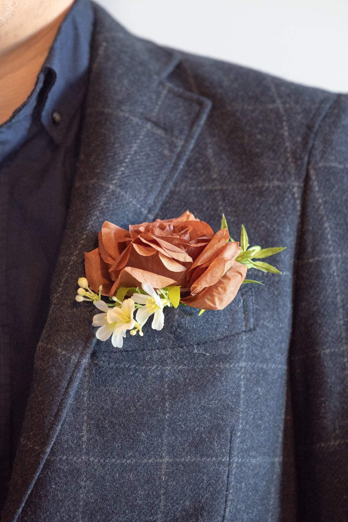 A close-up of a dark gray plaid suit jacket with a brown fabric rose boutonniere and small white flowers pinned to the chest pocket, worn over a navy blue shirt.